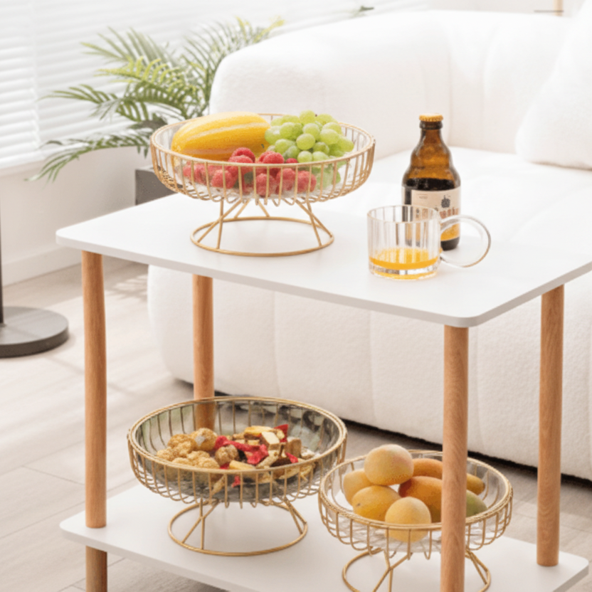 Small white table with gold wire fruit baskets containing fruits and a bottle on a light-colored floor.