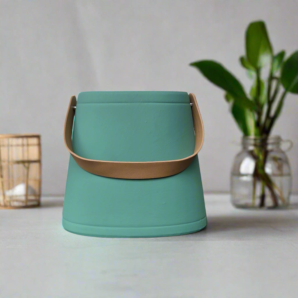 Green container with a brown handle on a light surface with a plant in the background