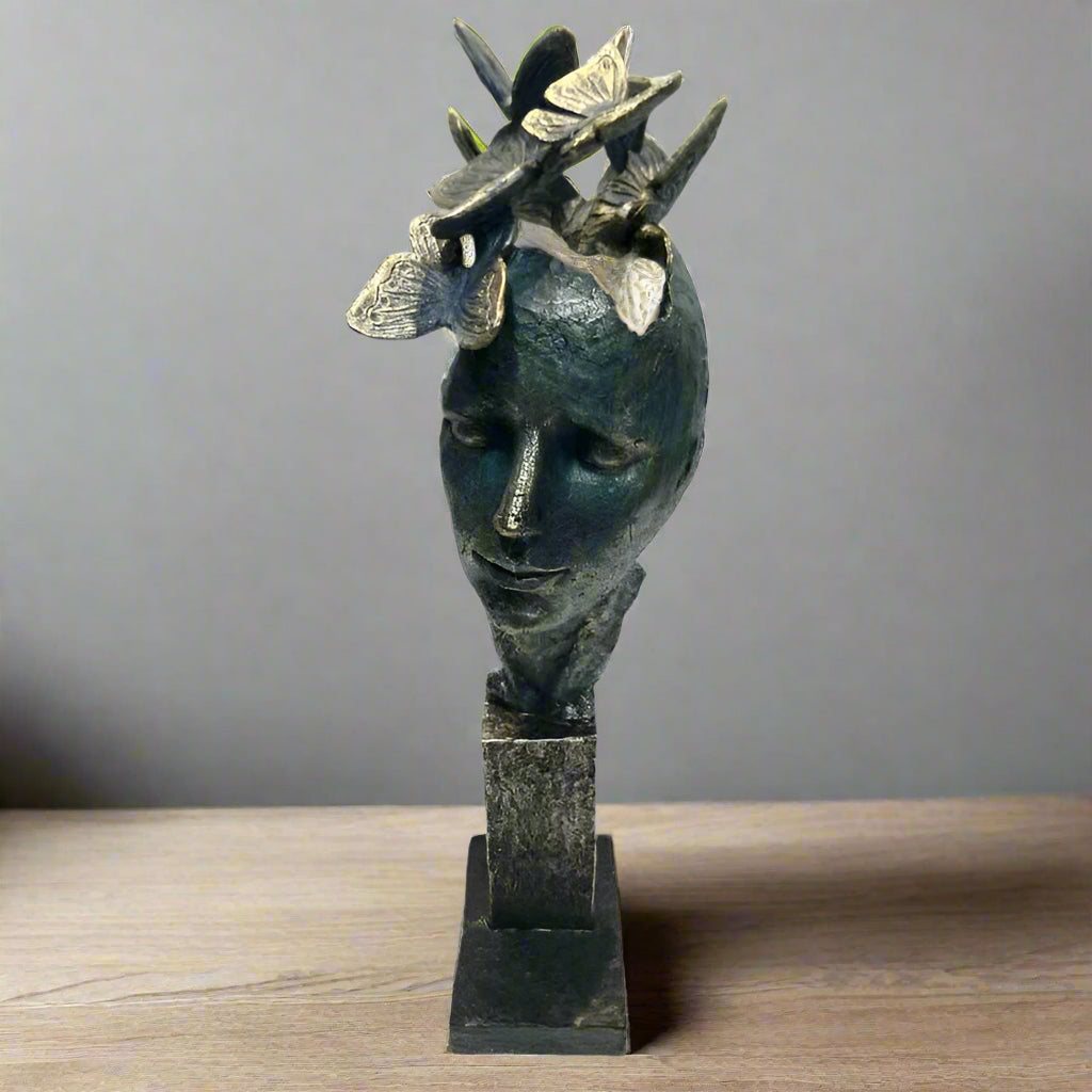 A stylized sculpture of a human head with butterflies around it, finished in dark, muted tones with a textured surface, displayed on a multi-tiered base.