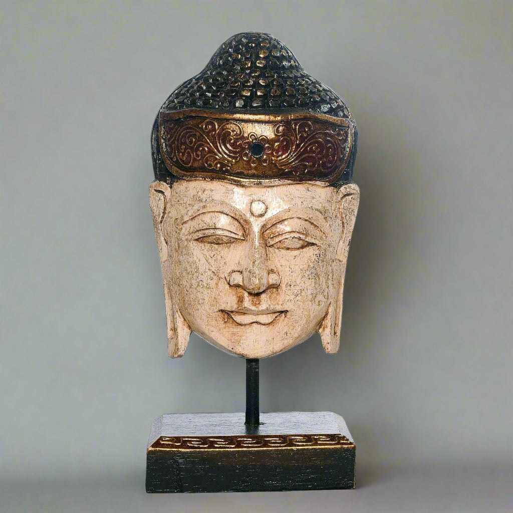 Buddha head sculpture with ornate headdress on a stand against a plain background