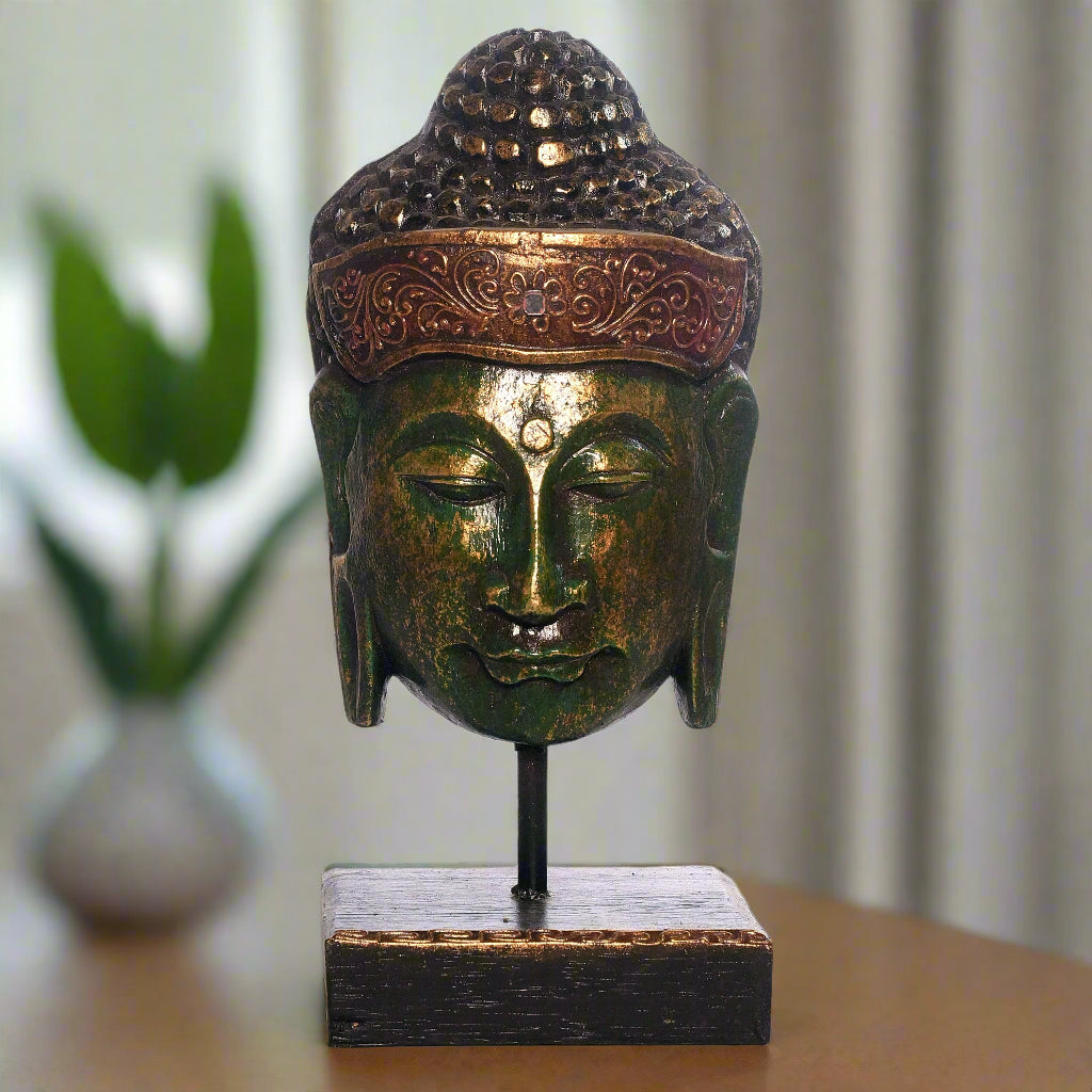 Bronze Buddha head sculpture on a stand with a blurred plant and curtain in the background