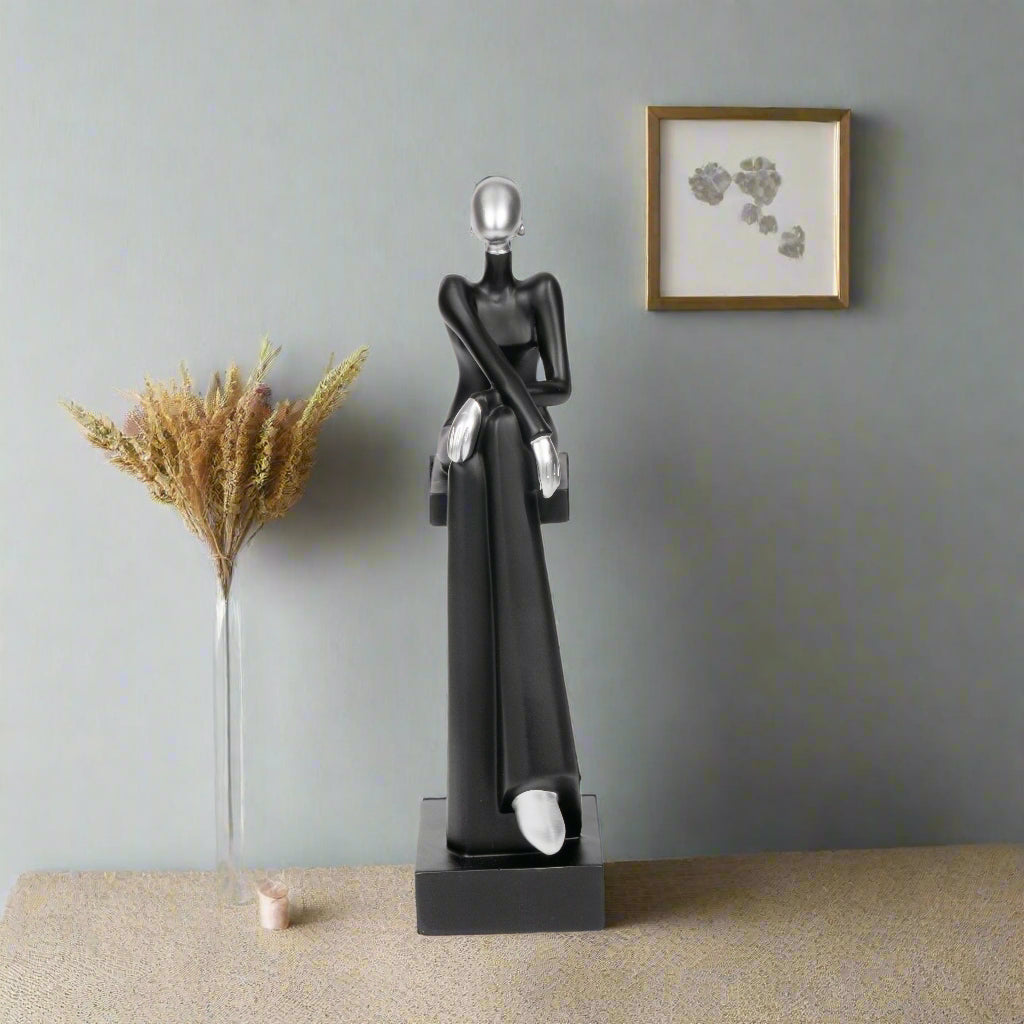 Black abstract sculpture on a beige surface with a vase of dried flowers and framed artwork in the background.