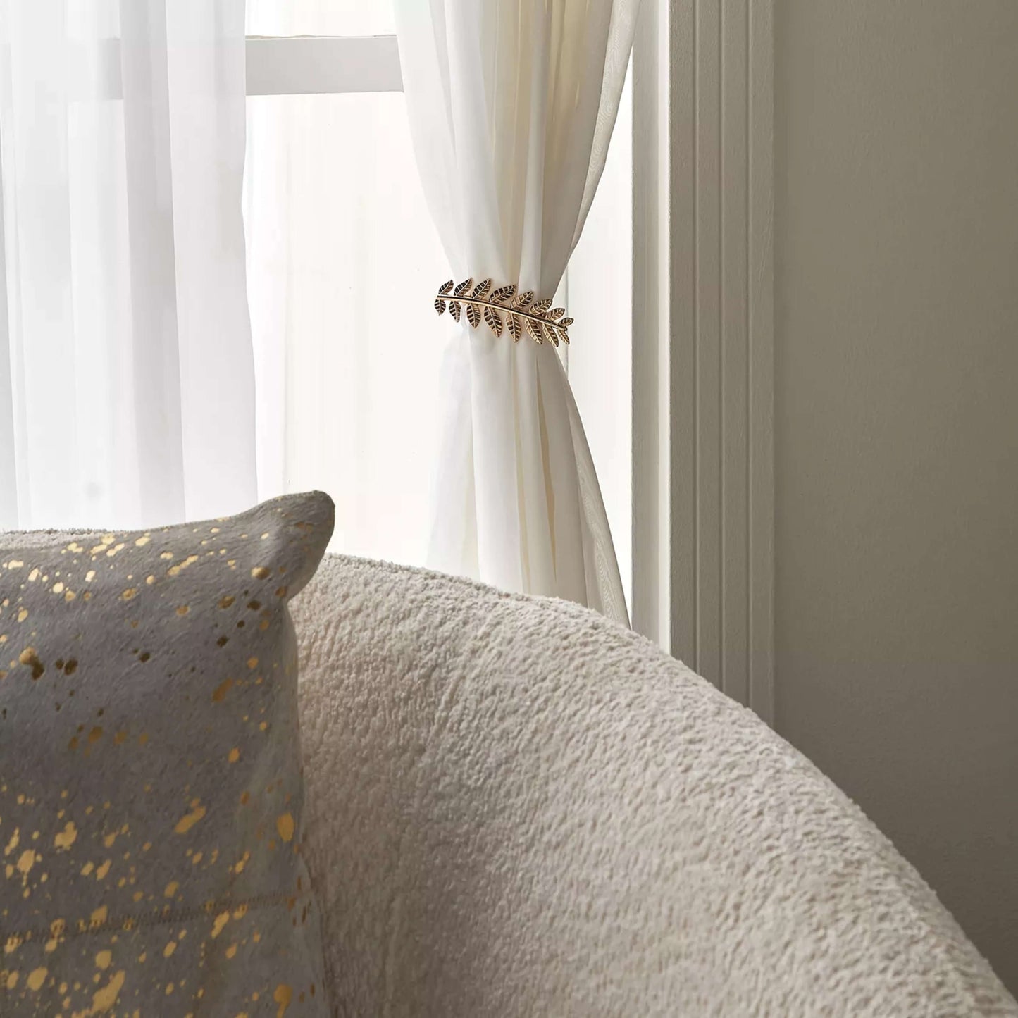 Decorative gold leaf curtain tieback on a white curtain with a textured gray sofa in the foreground.