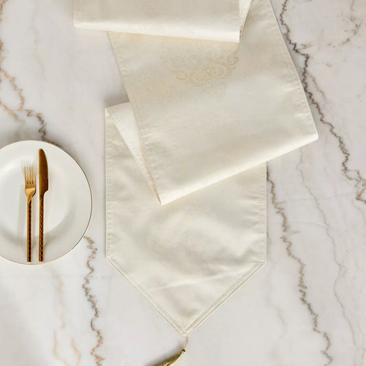 A white table runner with floral jacquard design and tassels on the sides, laid out on a marble-topped table with a golden fork and plate.