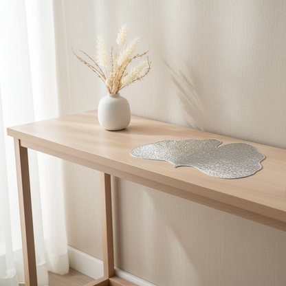Decorative leaf-shaped silver placemat on a wooden table with a vase of dried plants.