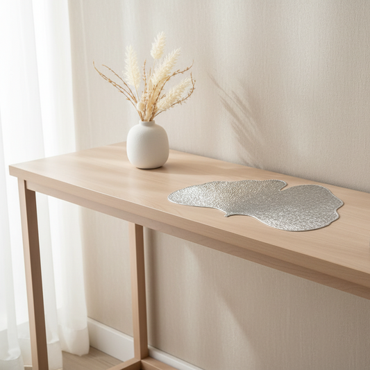 Decorative leaf-shaped silver placemat on a wooden table with a vase of dried plants.