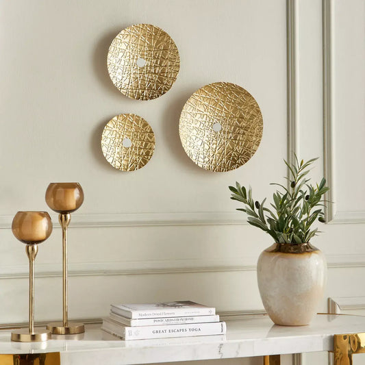 Decorative wall art with gold circular designs on a light gray wall, accompanied by gold candle holders and a plant on a marble surface.