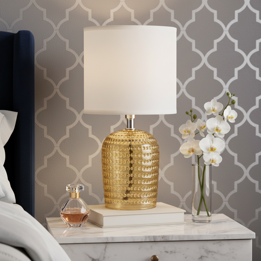 Gold lamp with white shade on a marble nightstand next to a vase of flowers and perfume bottle against a patterned wall.