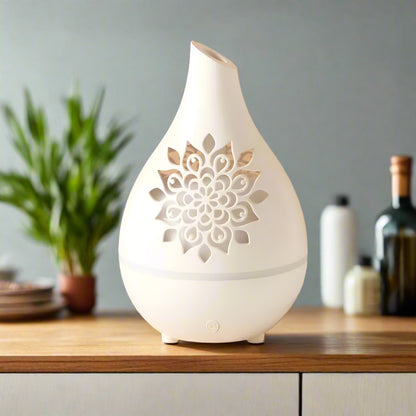 A white aroma diffuser with a vase-like design featuring a cutwork floral pattern, placed on a wooden surface with a glass vase of flowers in the background.