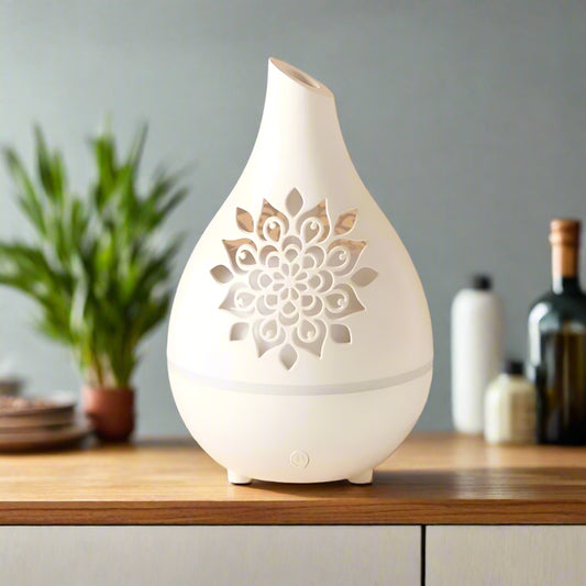 A white aroma diffuser with a vase-like design featuring a cutwork floral pattern, placed on a wooden surface with a glass vase of flowers in the background.