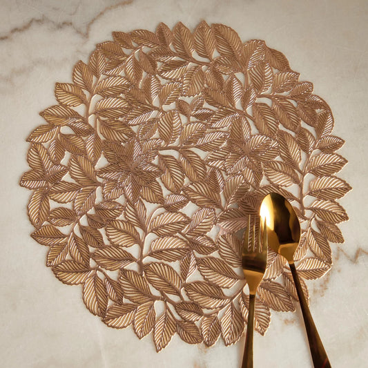 A gold-colored round placemat with a leaf pattern, displayed on a marble countertop alongside a spoon and a fork.