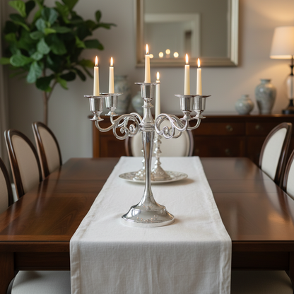 A silver three-arm candelabra with lit white taper candles is centered on a white table runner over a dark wood dining table, with matching chairs and a softly lit interior background.