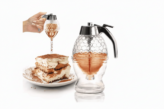 A clear glass syrup dispenser pitcher with a black metal spout and top, shown beside a hand holding the dispenser over a plate of tiramisu. The pitcher has a honeycomb pattern on the upper half and a clear lower half with liquid inside, on a white background.