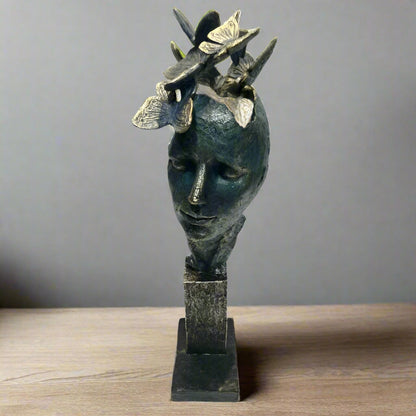 A stylized sculpture of a human head with butterflies around it, finished in dark, muted tones with a textured surface, displayed on a multi-tiered base.