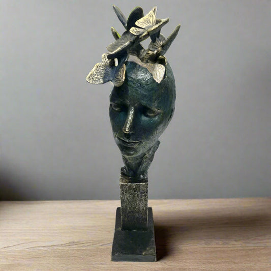 A stylized sculpture of a human head with butterflies around it, finished in dark, muted tones with a textured surface, displayed on a multi-tiered base.