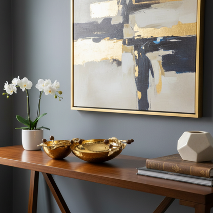 Decorative setup with gold bowls, a plant, books, and a abstract painting on a gray wall.