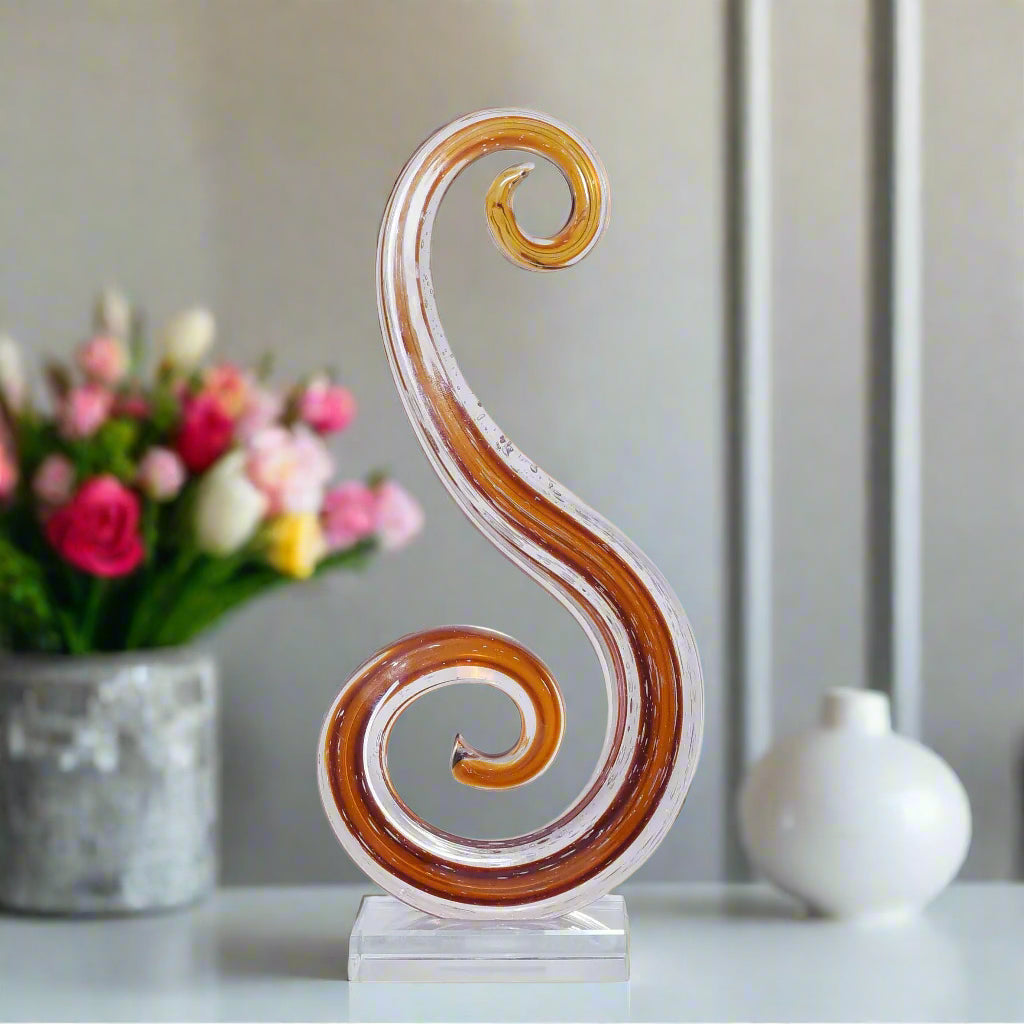 Decorative glass sculpture with swirling design on a wooden surface, with flowers in the background.