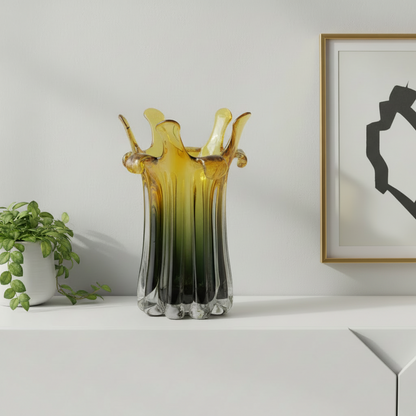 Decorative glass vase with gradient colors on a white surface next to a plant and framed artwork.
