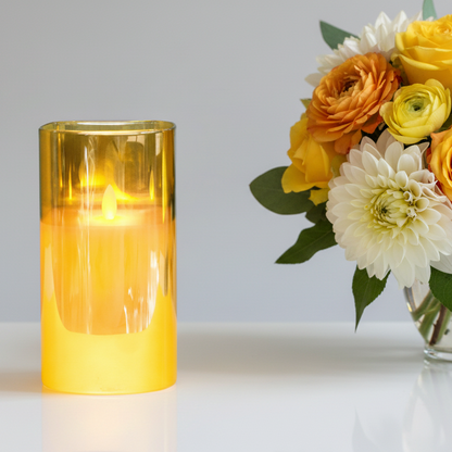 Yellow candle in a glass holder next to a bouquet of flowers on a white surface.