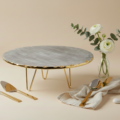 Round marble cake stand with gold base on a beige surface, accompanied by gold cutlery and flowers.