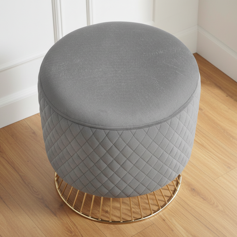 Gray quilted ottoman with a gold base on a wooden floor.