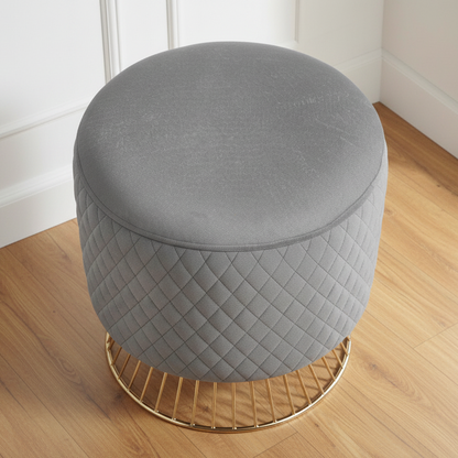 Gray quilted ottoman with a gold base on a wooden floor.